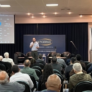 A imagem mostra um palestrante falando ao microfone em um auditório lotado. Do lado esquerdo da foto, há um telão. 