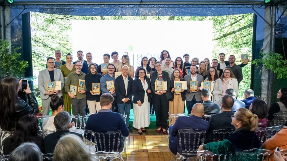 Uma foto de um Uma foto de um grupo grande de pessoas (cerca de 30, homens e mulheres) posando em um palco após receberem premiações. Eles estão de pé, sorrindo, e a maioria segura placas ou troféus retangulares com ilustrações. Eles são os vencedores do Prêmio Sema-Fepam de Jornalismo Ambiental 2025.
O palco é decorado com um grande painel de LED central que está totalmente iluminado com uma luz branca forte, e é emoldurado por painéis laterais que projetam imagens de folhagem verde, dando a impressão de natureza. Na frente do palco, há uma plateia sentada em cadeiras transparentes (de acrílico) e escuras. O ambiente é formal, mas com um toque de evento corporativo ou premiação.