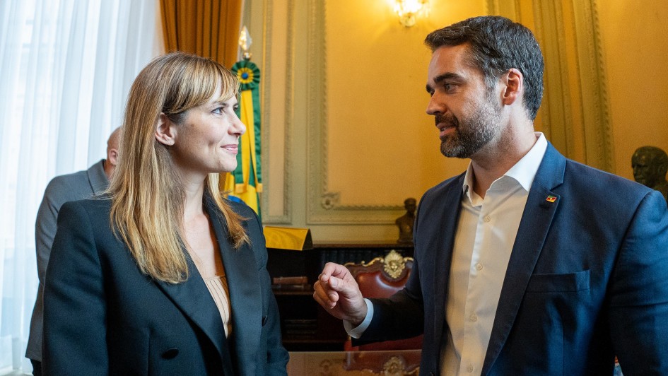 Uma mulher e um homem estão em primeiro plano em um ambiente interno. A mulher, à esquerda, é branca, tem cabelos loiros na altura dos ombros com franja e veste um blazer preto sobre uma blusa de cor clara. Ela está sorrindo e olhando para o homem. O homem, à direita, é branco, tem barba escura e veste um terno azul-marinho com um pin de bandeira no paletó e uma camisa social branca aberta no colarinho. Ele está olhando para a mulher e parece estar falando, gesticulando com a mão direita e segurando um pedaço de papel na esquerda. O fundo está um pouco desfocado e sugere uma sala formal, talvez um gabinete ou salão, com paredes em tons de amarelo ou dourado, cortinas pesadas, um busto ou estátua à direita, e o que parecem ser detalhes de uma bandeira ou estandarte (verde e amarelo) atrás da mulher.