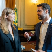 Uma mulher e um homem estão em primeiro plano em um ambiente interno. A mulher, à esquerda, é branca, tem cabelos loiros na altura dos ombros com franja e veste um blazer preto sobre uma blusa de cor clara. Ela está sorrindo e olhando para o homem. O homem, à direita, é branco, tem barba escura e veste um terno azul-marinho com um pin de bandeira no paletó e uma camisa social branca aberta no colarinho. Ele está olhando para a mulher e parece estar falando, gesticulando com a mão direita e segurando um pedaço de papel na esquerda. O fundo está um pouco desfocado e sugere uma sala formal, talvez um gabinete ou salão, com paredes em tons de amarelo ou dourado, cortinas pesadas, um busto ou estátua à direita, e o que parecem ser detalhes de uma bandeira ou estandarte (verde e amarelo) atrás da mulher.