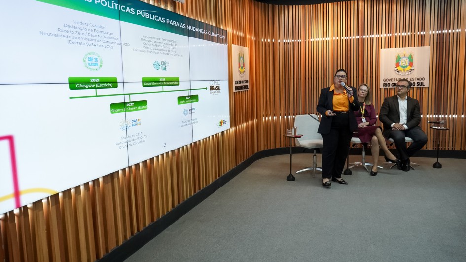 Aqui está uma descrição da imagem para fins de acessibilidade:A imagem exibe uma apresentação formal em um ambiente moderno, com paredes revestidas por painéis verticais de madeira ripada e piso acarpetado cinza.Plano Central e Pessoas:Uma mulher está de pé no centro-direita, falando em um microfone de mão e gesticulando levemente com a mão esquerda. Ela é branca, tem cabelos escuros presos e veste uma blusa laranja vibrante com calças sociais pretas. Ela é a mesma pessoa vista no slide anterior, provavelmente a Secretária Marjorie Kauffmann.Sentados à direita dela, em cadeiras de estofamento claro, estão duas outras pessoas. Uma mulher de casaco vinho/roxo está sentada à frente. Um homem de barba e terno escuro está sentado mais atrás, à extrema direita.Telão de Apresentação:Um grande telão horizontal na parede esquerda exibe o título "POLÍTICAS PÚBLICAS PARA AS MUDANÇAS CLIMÁTICAS".O conteúdo do slide é um fluxograma ou linha do tempo que detalha marcos e ações relacionadas a iniciativas climáticas, incluindo referências a eventos globais e legislação. Os itens visíveis incluem:Under 2 Coalition e Declaração de Edimburgo.Race to Zero / Race to Resilience.Neutralidade de emissões de $\text{CO}_2$ em 2050 (Decreto 56.341 de 2022).Datas e locais de COPs: 2021 Glasgow (Escócia), 2022 Sharm El Sheikh.2023 (Dubai).Menção a instrumentos jurídicos como a Lei Estadual do Clima e o Fundo Estadual do Clima.Fundo:À direita do telão, há um banner vertical com o brasão e o texto "GOVERNO DO ESTADO RIO GRANDE DO SUL".A cena retrata um painel de discussão ou apresentação sobre a agenda climática do estado.