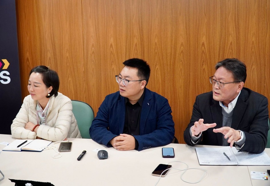 Três chineses estão sentadoss lado a lado em uma mesa de reuniões, conversando. A mulher à esquerda usa casaco claro, enquanto os dois homens à direita vestem casacos escuros e óculos. À frente deles há cadernos, celulares e cabos sobre a mesa.