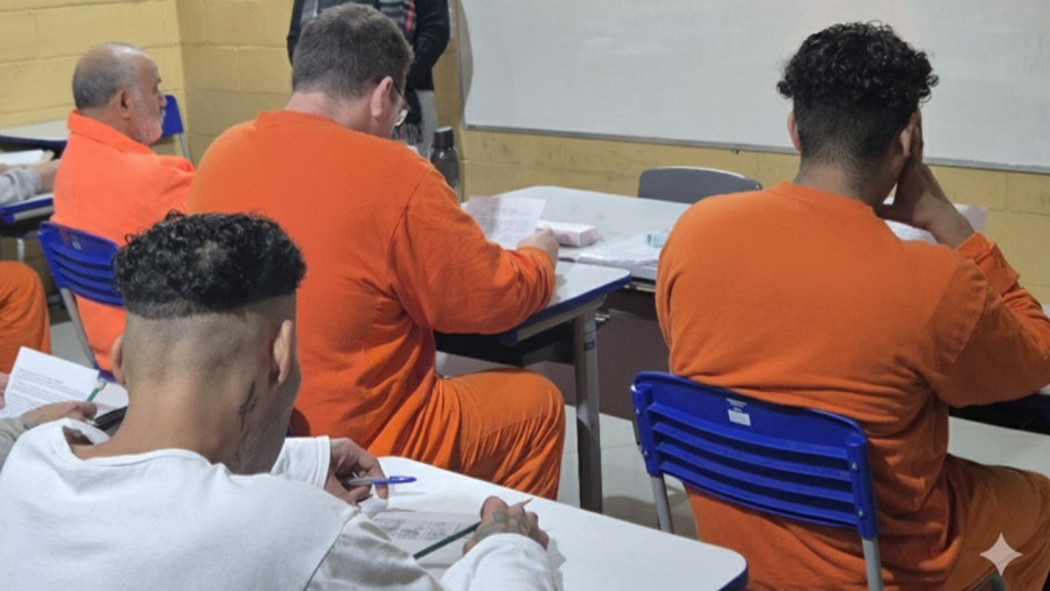 A imagem mostra quatro homens de costas, vestindo uniformes nas cores laranja e branco. Eles estão sentados em classes escolares, com papéis e canetas nas mãos.