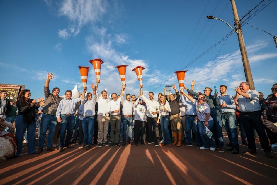 O governador Eduardo Leite aparece ao centro, junto a um grupo de pessoas, todas sorrindo e levantando cones de sinalização laranja no ar. O grupo está em uma estrada recém-pavimentada, sob céu azul, celebrando a inauguração da via.