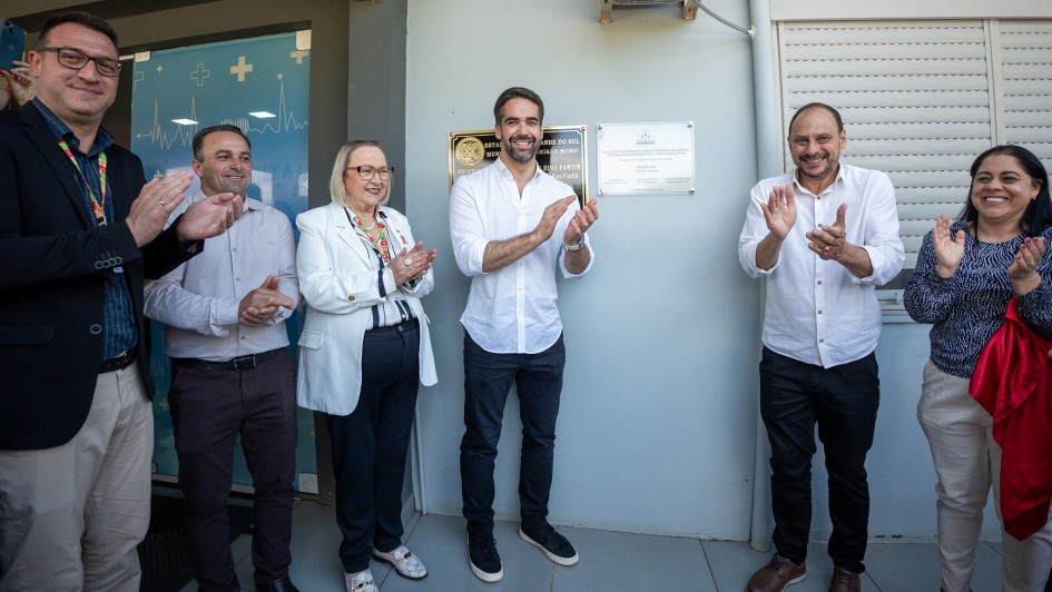 Governador Eduardo Leite inaugura reforma do pronto atendimento e entrega equipamentos a hospital em Mariano Moro com outras autoridades, em frente a uma placa. Eles sorriem e batem palmas.