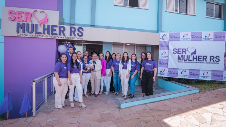 A foto mostra a fachada de um prédio pintado em tons de azul e lilás, com uma placa grande que diz “SER MULHER RS – Serviço Especializado de Referência à Saúde da Mulher”. Em frente à entrada, há um grupo de pessoas organizadas para uma foto coletiva, vestindo camisetas roxas.