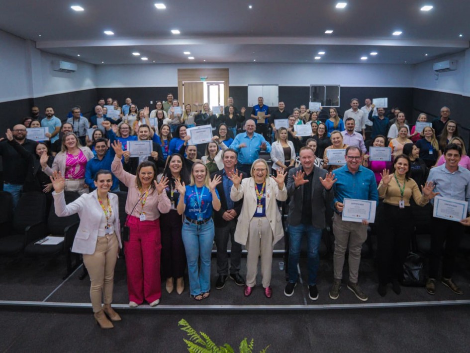 A foto mostra um auditório lotado, com pessoas organizadas em fileiras e um grupo maior posicionado à frente para foto. Várias pessoas seguram certificados.