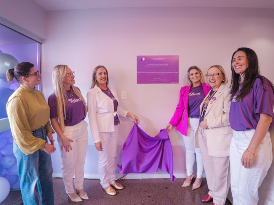 A foto mostra um grupo de pessoas reunidas em frente a uma placa de inauguração. No centro, duas pessoas seguram um tecido roxo parcialmente aberto, revelando uma placa comemorativa fixada na parede. A placa é retangular, com fundo roxo e texto branco, acompanhada do logotipo do programa SER Mulher RS.
