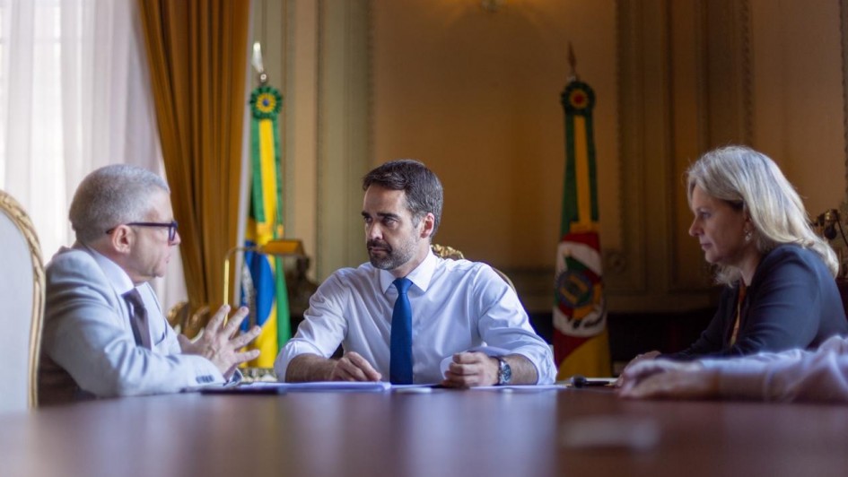 Foto de uma reunião num ambiente fechado. Dois homens e uma mulher aparecem sentados à mesa. Leite está na cabeceira, de frente para a câmera, olhando para o homem à esquerda, que fala.