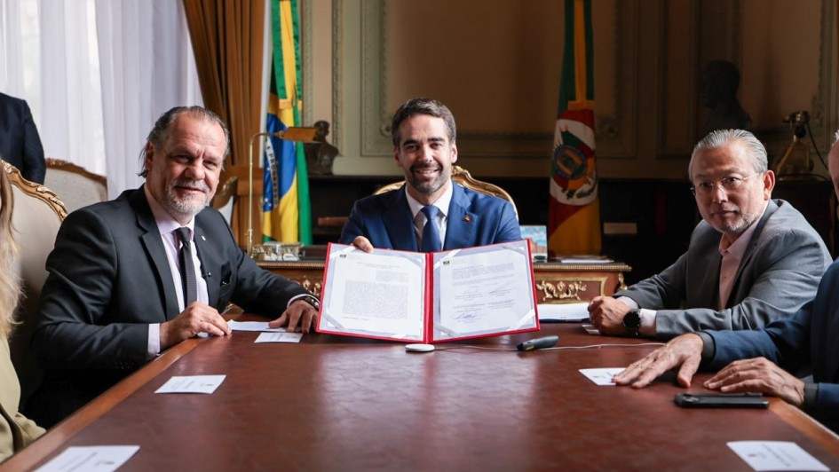 O governador Eduardo Leite aparece ao centro, segurando e exibindo um documento assinado, ao lado do presidente do Tribunal de Justiça do Rio Grande do Sul, desembargador Alberto Delgado Neto, à esquerda, e do secretário da Segurança Pública, coronel Mário Ikeda. A assinatura ocorre em sala com bandeiras do Brasil e do Estado ao fundo.