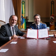 O governador Eduardo Leite aparece ao centro, segurando e exibindo um documento assinado, ao lado do presidente do Tribunal de Justiça do Rio Grande do Sul, desembargador Alberto Delgado Neto, à esquerda, e do secretário da Segurança Pública, coronel Mário Ikeda. A assinatura ocorre em sala com bandeiras do Brasil e do Estado ao fundo.