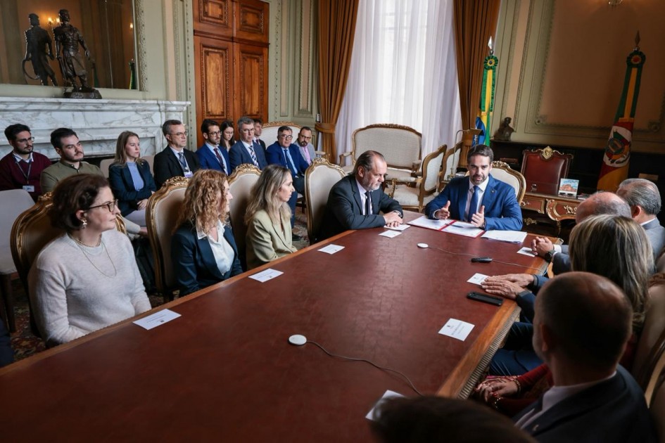 O governador Eduardo Leite aparece à direita da mesa, falando durante a cerimônia de assinatura. À sua frente estão o presidente do Tribunal de Justiça, desembargador Alberto Delgado Neto, e demais autoridades e servidores, incluindo o secretário da Segurança Pública, coronel Mário Ikeda, e o chefe de Polícia, delegado Heraldo Chaves Guerreiro, sentados ao redor da mesa.