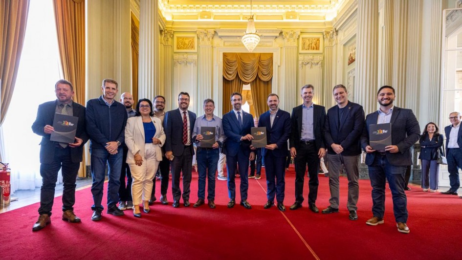 O governador Eduardo Leite aparece ao centro, de terno azul e gravata, posando para foto com um grupo de pessoas no no Palácio Piratini. O grupo está disposto em fileira sobre um tapete vermelho, segurando pastas pretas com o brasão do Governo do Estado.
