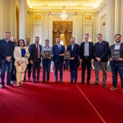 O governador Eduardo Leite aparece ao centro, de terno azul e gravata, posando para foto com um grupo de pessoas no no Palácio Piratini. O grupo está disposto em fileira sobre um tapete vermelho, segurando pastas pretas com o brasão do Governo do Estado.