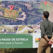 O governador Eduardo Leite (à esquerda, de camisa verde) observa um grande painel com o mapa e o plano urbanístico do Novo Passo de Estrela, acompanhado por um técnico que usa capacete branco e colete azul com o brasão do Estado. O painel mostra o traçado do novo bairro, áreas de acesso e o slogan “Um olhar para o futuro”.