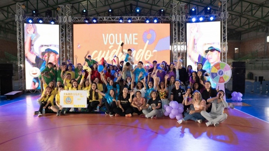 A imagem mostra um grupo numeroso de jovens em um palco sorrindo e com os braços levantados. Alguns estão em pé, e outros estão sentados, posando para a foto. 