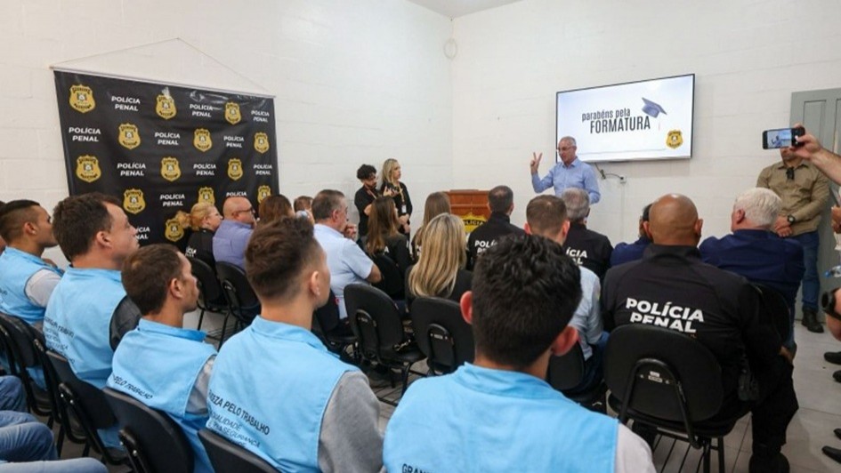 A imagem mostra um grupo de pessoas de costas, sentadas em cadeiras alinhadas. Na frente deles, está o secretário de Sistemas Penal e Socioeducativo, falando, enquanto no fundo há uma tela onde se lê "Parabéns pela formatura". Do lado esquerdo, há a marca da Polícia Penal em um banner. 