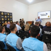 A imagem mostra um grupo de pessoas de costas, sentadas em cadeiras alinhadas. Na frente deles, está o secretário de Sistemas Penal e Socioeducativo, falando, enquanto no fundo há uma tela onde se lê "Parabéns pela formatura". Do lado esquerdo, há a marca da Polícia Penal em um banner. 