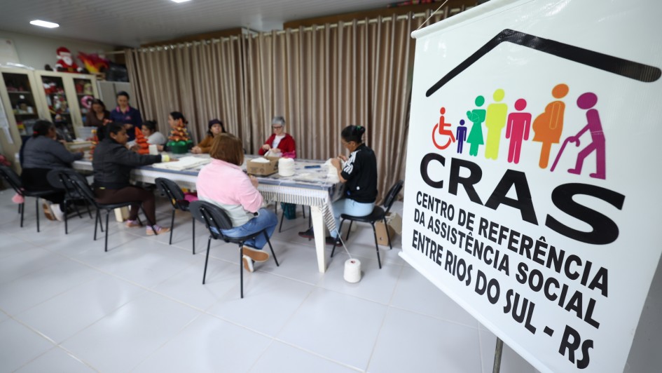 Foto mostra grupo de oito mulheres sentadas ao redor de uma grande mesa retangular e praticando crochê