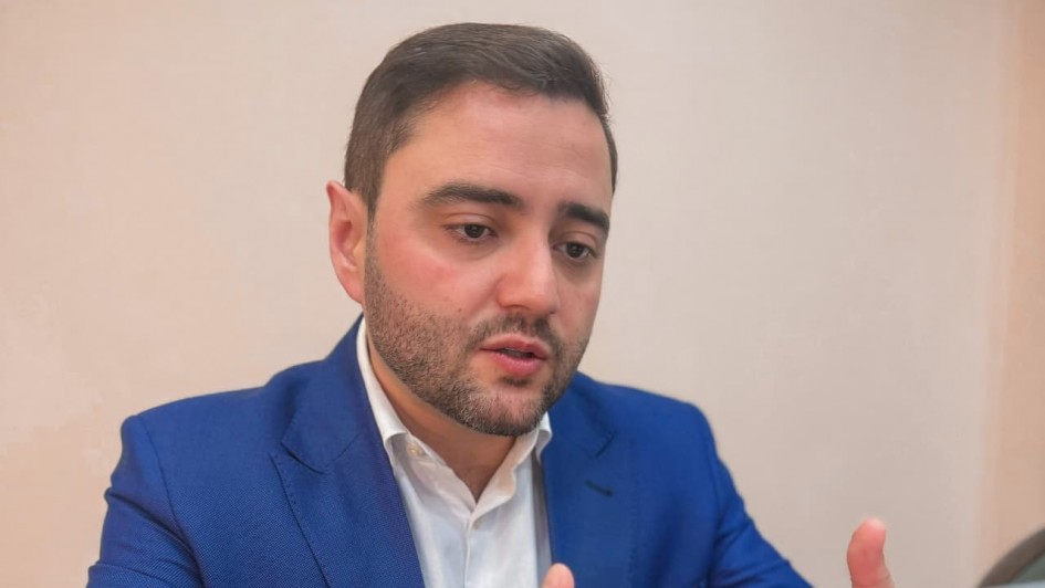 Uma foto em plano médio de um homem de pele clara, cabelo escuro e barba curta, vestido com um blazer azul vibrante sobre uma camisa social branca. Ele está sentado em uma mesa e gesticula com as mãos enquanto olha para frente, possivelmente para uma tela de notebook parcialmente visível no canto inferior direito. O fundo é de cor bege claro, liso e discreto. A iluminação é suave, destacando o rosto e o blazer do homem.