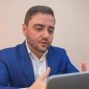 Uma foto em plano médio de um homem de pele clara, cabelo escuro e barba curta, vestido com um blazer azul vibrante sobre uma camisa social branca. Ele está sentado em uma mesa e gesticula com as mãos enquanto olha para frente, possivelmente para uma tela de notebook parcialmente visível no canto inferior direito. O fundo é de cor bege claro, liso e discreto. A iluminação é suave, destacando o rosto e o blazer do homem.