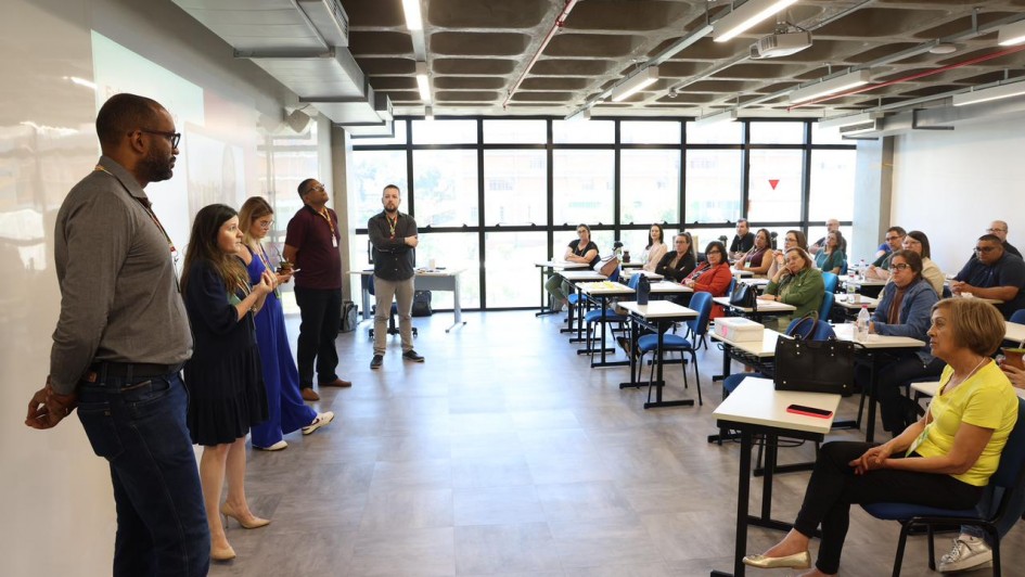 Foto exibe sala de aula com palestrantes, de pé à esquerda, e ouvintes participantes da formação sentados nas classes.