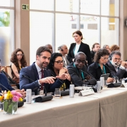 Foto de Leite sentado ao lado de várias pessoas numa mesa comprida, em uma conferência. O ambiente é formal. Leite fala ao microfone e gesticula com as mãos.