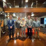 A imagem mostra cinco homens em pé, posando para a foto. No lado esquerdo da imagem, há uma fotografia em um cavalete. No lado direito, há um painel com uma apresentação sobre os 400 anos das Missões. 