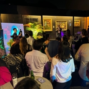 Foto mostra o lançamento da programação da Noite dos Museus, com público reunido em frente a um palco interno decorado com plantas e obras de arte expostas nas paredes. A iluminação em tons quentes e o painel com o slogan “A arte desperta” compõem o cenário, enquanto um orador faz pronunciamento diante da plateia atenta.