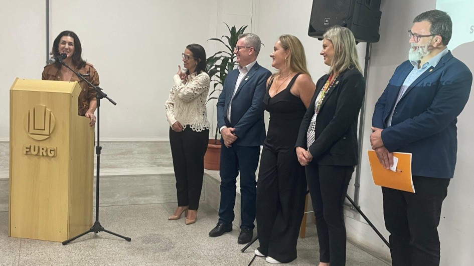 Foto mostra secretária discursando num púlpito com a logo da Furg. Ao lado dela, em pé, estão cinco pessoas, dois homens e três mulheres, que a observam atentamente.