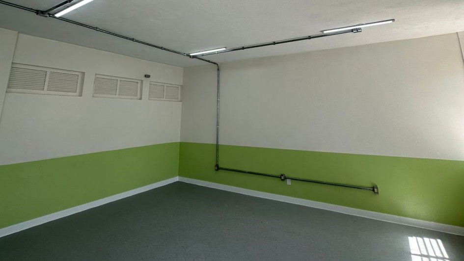 A imagem mostra uma sala de aula vazia, com as paredes nas cores branca e verde. O piso é cinza e há lâmpadas iluminando o ambiente. 