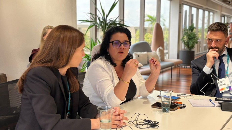 Foto de três pessoas sentadas junto a uma mesa de reunião, num ambiente formal fechado. Ao centro, Marjorie aparece de blusa branca falando e gesticulando com as mãos.
