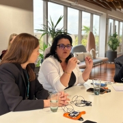 Foto de três pessoas sentadas junto a uma mesa de reunião, num ambiente formal fechado. Ao centro, Marjorie aparece de blusa branca falando e gesticulando com as mãos.