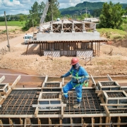 Três trabalhadores com uniformes azuis e capacetes vermelhos estão sobre uma estrutura de concreto e aço em construção de uma ponte. Eles usam cintos de segurança e estão posicionados sobre vigas e armações metálicas. Ao fundo, há outra parte da ponte em fase mais avançada, com colunas e lajes já concretadas. 