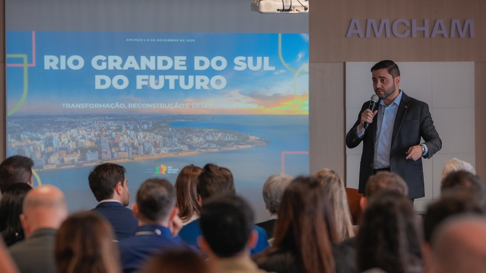 Uma foto interna em close-up de uma palestra ou evento em um auditório ou sala de conferências. Um homem (Gabriel Souza), no lado direito, está de pé no palco, vestindo um blazer escuro sobre uma camisa social azul-clara, e falando em um microfone que segura com a mão direita.

Atrás e à esquerda dele, há uma grande tela de projeção. O slide exibido na tela é dominado pela imagem de uma paisagem urbana à beira-mar (possivelmente Porto Alegre). No topo do slide, o texto em letras grandes e coloridas diz "RIO GRANDE DO SUL DO FUTURO" e, abaixo, "TRANSFORMAÇÃO, RECONSTRUÇÃO E DESENVOLVIMENTO".

Em primeiro plano, a parte superior da cabeça e dos ombros de vários membros da audiência, que estão de costas para a câmera, preenche o terço inferior da imagem. No canto superior direito, há uma placa com o nome "AMCHAM". A iluminação é focada no palestrante e no slide.