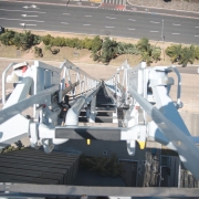 A imagem apresenta uma perspectiva vertical a partir do topo de uma autoescada mecânica, mostrando a estrutura metálica da escada em primeiro plano. Abaixo, é possível ver uma avenida larga com faixas de trânsito e algumas árvores.