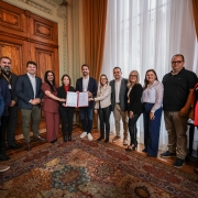 Uma foto colorida, tirada em ambiente interno que parece ser um gabinete ou sala de reuniões formal, possivelmente em um palácio de governo, devido à arquitetura e decoração. Um grupo de doze pessoas, composto por homens e mulheres, está posicionado no centro da sala, em pé sobre um tapete estampado.

No centro do grupo, um homem (aparentemente o governador) e uma mulher estão segurando um documento (possivelmente um ato de autorização ou convênio), indicando uma assinatura ou entrega formal.

Pessoas: Estão vestidas com trajes sociais ou casuais de negócios. O homem no centro-direita (governador) veste blazer e camisa polo. Outros homens usam ternos, blazers ou camisas sociais. As mulheres usam blazers, calças sociais e blusas.

Cenário: A sala tem um alto pé-direito. Ao fundo, uma grande porta de madeira escura com painéis esculpidos ocupa a parede central. À direita, janelas altas estão cobertas por cortinas longas e pesadas, de cor bege ou dourada, que permitem a entrada de luz natural. O piso é coberto por um grande tapete oriental, ricamente estampado em tons de bordô, azul-marinho e bege. A iluminação é clara e uniforme.

A pose é formal, com todos olhando para a câmera e sorrindo, sugerindo um momento de celebração ou registro de um acordo importante.