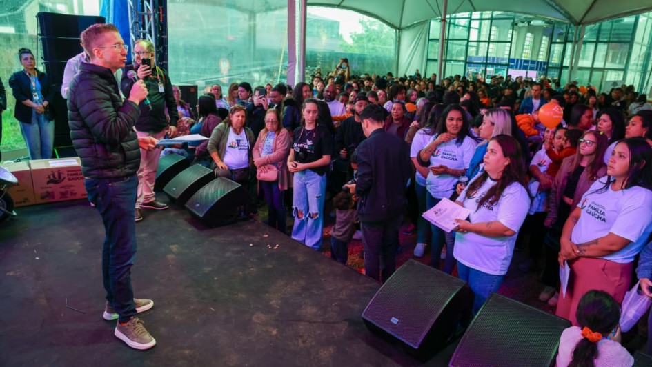 Foto de Beto, de pé à esquerda, falando ao microfone numa tenda, com a plateia de pé à direita do quadro.