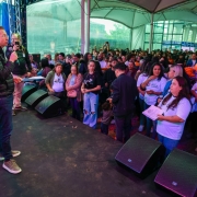 Foto de Beto, de pé à esquerda, falando ao microfone numa tenda, com a plateia de pé à direita do quadro.