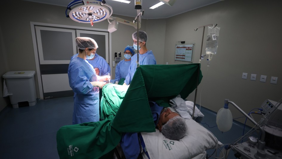 Equipe médica realiza uma cirurgia de joelho em um centro cirúrgico. A cena mostra profissionais de saúde vestidos com aventais e toucas esterilizadas, concentrados no procedimento sob iluminação cirúrgica. O paciente está deitado, anestesiado e coberto por panos verdes, em ambiente hospitalar moderno e bem iluminado.