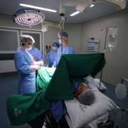 Equipe médica realiza uma cirurgia de joelho em um centro cirúrgico. A cena mostra profissionais de saúde vestidos com aventais e toucas esterilizadas, concentrados no procedimento sob iluminação cirúrgica. O paciente está deitado, anestesiado e coberto por panos verdes, em ambiente hospitalar moderno e bem iluminado.