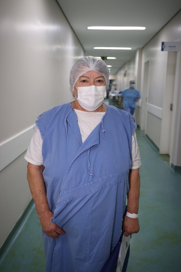 Mulher idosa, vestida com avental azul e máscara, é retratada em um corredor hospitalar. A paciente olha para a câmera com expressão tranquila.