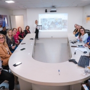 Uma foto colorida mostra um grupo de pessoas em uma sala de reuniões. A sala é bem iluminada e possui paredes claras.

Mesa: Há uma grande mesa de reunião em formato de 'U' ou ferradura, com tampo na cor bege ou creme. Vários participantes estão sentados ao longo das laterais curvas da mesa.

Participantes: A maioria dos presentes são mulheres. Elas estão vestindo roupas casuais de negócios em cores variadas. No canto inferior esquerdo, um homem de terno cinza e óculos está sentado mais próximo da câmera. Outras pessoas estão sentadas no lado esquerdo da mesa e no lado curvo (ao fundo).

Apresentadora: No centro da parte curva da mesa, uma mulher de blusa branca está em pé, segurando o que parece ser um documento ou tablet. Ela está de frente para o grupo.

Projeção: Atrás da apresentadora, há um projetor exibindo uma tela que mostra várias imagens em miniatura (possivelmente de outras reuniões ou pessoas).

Detalhes da Sala: No canto superior esquerdo da parede, há um cartaz parcialmente visível com texto relacionado a "CORONAVÍRUS" e "RS Contra Gripe".

A imagem parece registrar um evento formal, como uma reunião ou a assinatura de um convênio, dado o título do arquivo