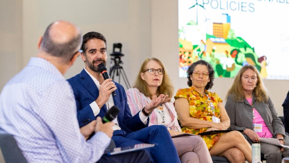 A imagem mostra o governador Eduardo Leite falando ao microfone. Ao lado dele, há outras quatro pessoas sentadas, compondo uma mesa-redonda. Ao fundo, há um painel com uma apresentação. 