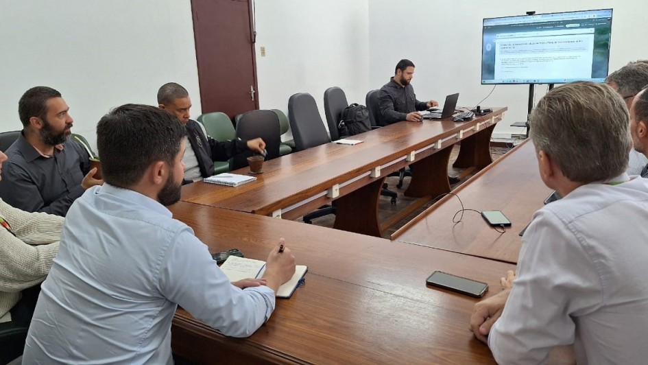 Uma foto colorida registra uma reunião em uma sala de conferências. A imagem é cortada de forma a mostrar a maioria dos participantes e a mesa.

Mesa: Uma grande mesa de reunião de madeira escura em formato de 'U' ou retangular alongado domina o centro.

Participantes: Cerca de oito pessoas (a maioria homens) estão sentadas em volta da mesa.

Primeiro Plano: No lado esquerdo, um homem de camisa social azul clara está de costas para a câmera, escrevendo em um bloco de notas.

Lado Esquerdo: Uma mulher de suéter de malha claro está sentada ao lado do homem de camisa azul, parcialmente visível. Outros homens estão sentados nesse lado da mesa. Um deles parece estar segurando uma cuia de chimarrão.

Ao Fundo: Um homem de camisa escura está sentado ao centro do fundo da mesa, trabalhando em um laptop.

Primeiro Plano Direito: Vários homens estão sentados de costas para a câmera no lado direito da mesa, com um deles parcialmente visível.

Tela: À direita do homem do laptop, uma tela de televisão ou monitor está montada na parede. Ela exibe um documento digital ou formulário com texto.

Detalhes da Sala: A sala tem paredes claras e algumas cadeiras de escritório pretas com encosto alto estão visíveis no fundo. Há alguns itens como celulares e cadernos sobre a mesa.

A imagem sugere uma discussão ou apresentação de trabalho, possivelmente relacionada ao formulário de adesão mencionado no nome do arquivo
