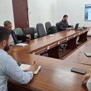 Uma foto colorida registra uma reunião em uma sala de conferências. A imagem é cortada de forma a mostrar a maioria dos participantes e a mesa.

Mesa: Uma grande mesa de reunião de madeira escura em formato de 'U' ou retangular alongado domina o centro.

Participantes: Cerca de oito pessoas (a maioria homens) estão sentadas em volta da mesa.

Primeiro Plano: No lado esquerdo, um homem de camisa social azul clara está de costas para a câmera, escrevendo em um bloco de notas.

Lado Esquerdo: Uma mulher de suéter de malha claro está sentada ao lado do homem de camisa azul, parcialmente visível. Outros homens estão sentados nesse lado da mesa. Um deles parece estar segurando uma cuia de chimarrão.

Ao Fundo: Um homem de camisa escura está sentado ao centro do fundo da mesa, trabalhando em um laptop.

Primeiro Plano Direito: Vários homens estão sentados de costas para a câmera no lado direito da mesa, com um deles parcialmente visível.

Tela: À direita do homem do laptop, uma tela de televisão ou monitor está montada na parede. Ela exibe um documento digital ou formulário com texto.

Detalhes da Sala: A sala tem paredes claras e algumas cadeiras de escritório pretas com encosto alto estão visíveis no fundo. Há alguns itens como celulares e cadernos sobre a mesa.

A imagem sugere uma discussão ou apresentação de trabalho, possivelmente relacionada ao formulário de adesão mencionado no nome do arquivo