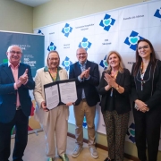 A imagem mostra cinco pessoas posando lado a lado. A secretária da Saúde segura e exibe um documento assinado.