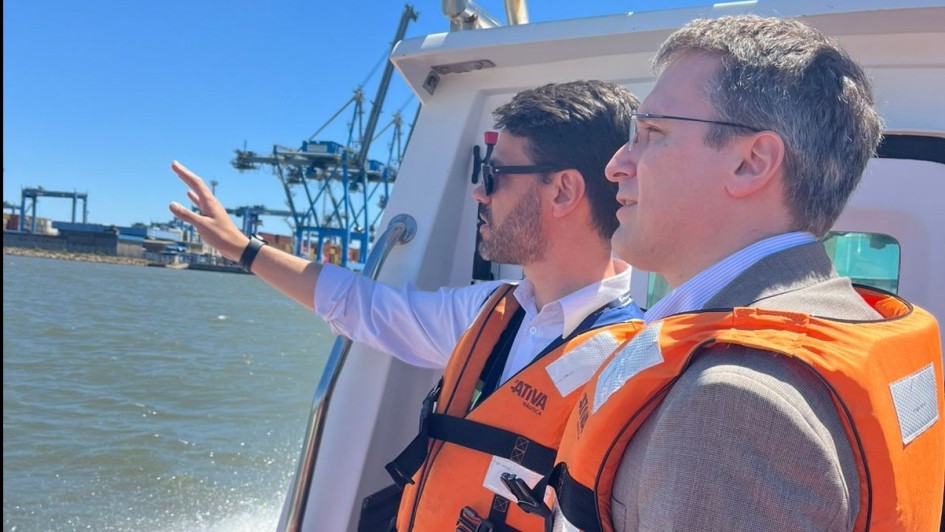 Foto de dois homens a c&eacute;u aberto, navegando num barco. Eles aparecem de lado, trajando coletes laranjas de boiar. Um deles faz um gesto com o bra&ccedil;o direito erguido na dire&ccedil;&atilde;o de um lugar fora do quadro, para o qual os dois olham.