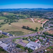 A imagem mostra uma vista aérea de uma área semiurbana. Em primeiro plano, há uma rodovia com veículos e prédios industriais e comerciais ao redor. Mais ao fundo, aparecem grandes áreas verdes e campos, com algumas construções espalhadas. À direita, há um bairro com muitas casas e ruas organizadas. No horizonte, vê-se colinas e vegetação sob um céu azul claro.
