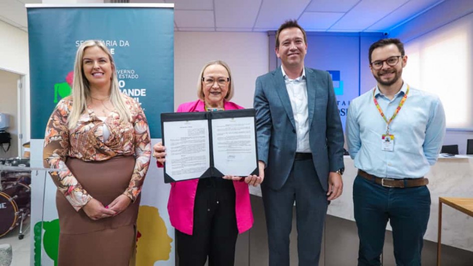A imagem mostra quatro pessoas adultas em pé, lado a lado, em um ambiente interno, posando para uma foto oficial após a assinatura de um documento.

Pessoas (da esquerda para a direita):

Mulher: Veste uma blusa estampada em tons de pêssego e marrom sobre uma saia marrom-clara. Ela é loira, com um sorriso voltado para a câmera.

Mulher (Central): Veste uma jaqueta de cor rosa choque sobre calças pretas. Ela usa óculos e segura um documento dobrado (possivelmente um contrato ou convênio) em uma pasta preta, apresentando o papel aberto para a câmera.

Homem: Veste um terno cinza escuro sobre uma camisa branca sem gravata. Ele é moreno, com cabelo curto, sorrindo para a câmera.

Homem (Extrema Direita): Veste uma camisa social azul-clara e calças jeans escuras. Ele usa óculos e um crachá pendurado em um cordão laranja/vermelho.

Fundo:

No canto esquerdo, há um banner vertical turquesa (ou azul esverdeado) com texto parcial visível que inclui as palavras "SECRETARIA DA", "GOVERNO ESTADO" e "GRANDE".

No centro e à direita, a parede de fundo é clara, com um segundo banner ou painel no canto superior direito que mostra parte de um logotipo azul e branco, possivelmente institucional ("TAI S I").

No canto inferior esquerdo, parte de um kit de bateria (tambores vermelhos e brancos) é visível.